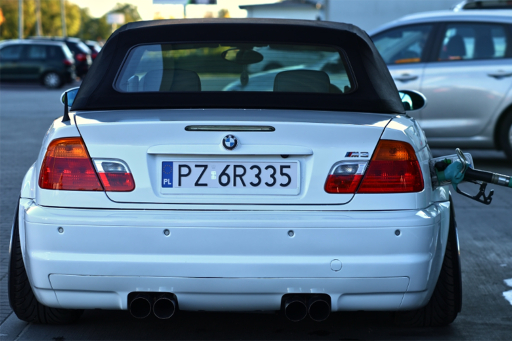 Biały kabriolet BMW widziany od tyłu na stacji benzynowej, z pistoletem dystrybutora włożonym do wlewu paliwa.