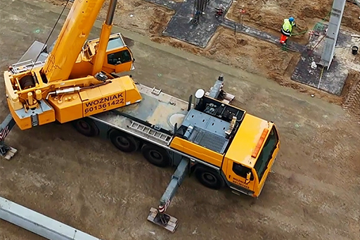 Dźwig na placu budowy, z pracownikami w kamizelkach, przy metalowych konstrukcjach.