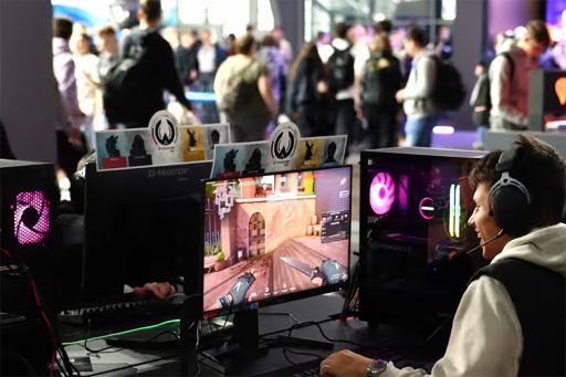 Gracz w słuchawkach siedzący przy komputerze z kolorowo podświetlonym PC i monitorem z grą FPS, na tle tłumu ludzi na targach gamingowych.