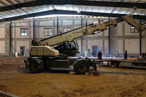 Samochód z teleskopowym dźwigiem pracujący wewnątrz dużej hali w budowie, na piaszczystym podłożu, z kilkoma pracownikami w tle.