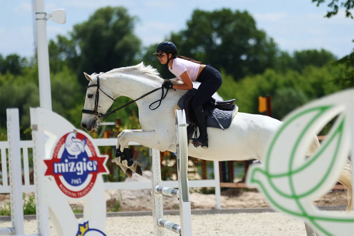 Zawodniczka w kasku na białym koniu w trakcie skoku przez przeszkodę na parkurze, na tle zielonych drzew.