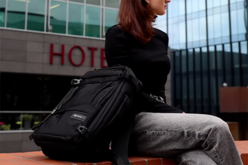 Młoda kobieta siedząca na murku z dużym czarnym plecakiem przy boku na tle nowoczesnego, przeszklonego budynku.