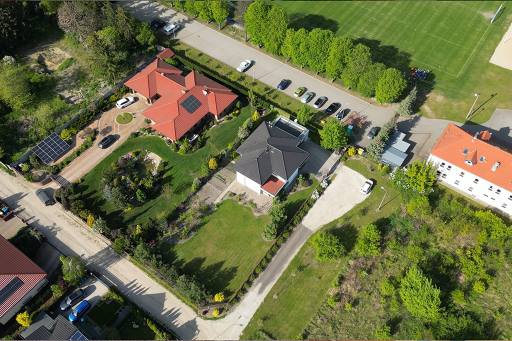 Osiedle domów jednorodzinnych z zadbanymi ogrodami widziane z lotu ptaka, z drogą pełną zaparkowanych samochodów i boiskiem sportowym po prawej stronie.