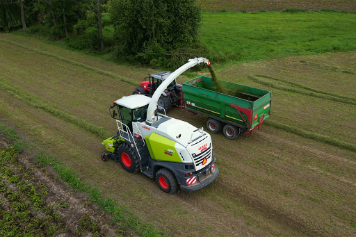 Samobieżna sieczkarnia polowa z białą kabiną napełnia zieloną przyczepę ciągniętą przez czerwony traktor na łące widzianej z góry.