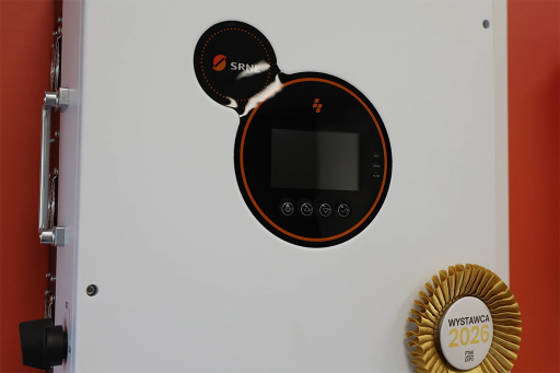 Białe urządzenie elektryczne z okrągłym czarnym panelem sterowania na pomarańczowej ścianie, ozdobione złotą rozetką nagrody w prawym dolnym rogu.